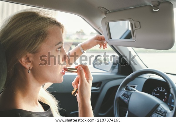 Beautiful woman with blond\
hair driving a car paints her lips with lipstick. The woman driving\
making make-up looking in the Decorative makeup cosmetic mirror for\
sun visor