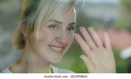 Beautiful Woman With Blond Hair Watching The View By The Window. She Salutes His Neighbors Waving. There Is A Reflection Of The Scenery In The Glass.