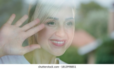 Beautiful Woman With Blond Hair Watching The View By The Window. She Salutes His Neighbors Waving. There Is A Reflection Of The Scenery In The Glass.