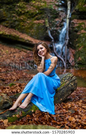 A beautiful woman in blue dress near a waterfall in autumn day