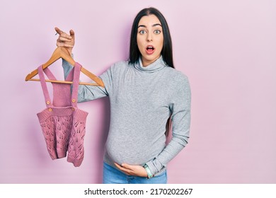 Beautiful Woman With Blue Eyes Expecting A Baby Holding Baby Clothes Afraid And Shocked With Surprise And Amazed Expression, Fear And Excited Face. 