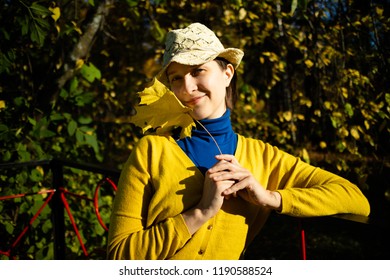 Beautiful Woman In Blue Polo Neck And Yellow Cardigan And A Hat Is Holding Yellow Maple Leaf In Autumn Park