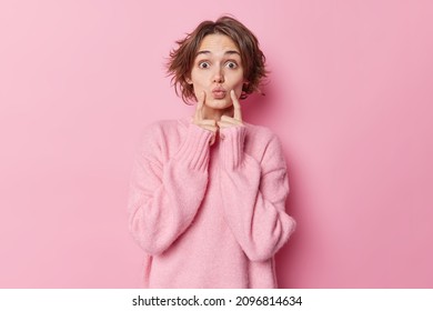 Beautiful Woman With Bob Hairstyle Keeps Lips Rounded Index Fingers On Cheeks Has Romantic Look Awaits For Kiss Wears Warm Cashmere Jumper Isolated Over Pink Background. Face Expressions Concept