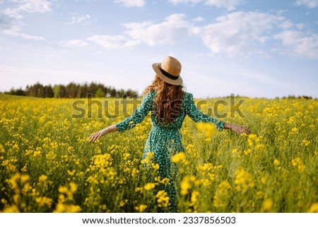 Beautiful woman in bright dress and elegant hat walks and has fun in rapeseed field. Smiling female tourist walking through flowering field, touching yellow flowers. Nature, rest. Summer landscape.