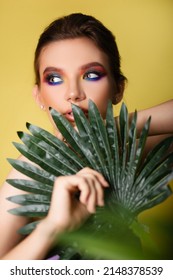 Beautiful Woman With Bright Makeup On A Yellow Background In A Purple Sportswear With A Palm Leaf. Girl On Vacation. Photo In The Style Of The 80s