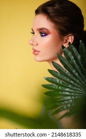 Beautiful Woman With Bright Makeup On A Yellow Background In A Purple Sportswear With A Palm Leaf. Girl On Vacation. Photo In The Style Of The 80s