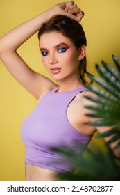 Beautiful Woman With Bright Makeup On A Yellow Background In A Purple Sportswear With A Palm Leaf. Girl On Vacation. Photo In The Style Of The 80s