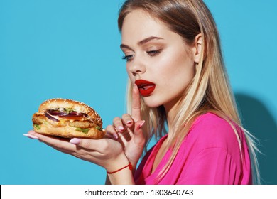 beautiful woman with bright makeup in a pink shirt looks at a hamburger on a blue pony isolated - Shutterstock ID 1303640743