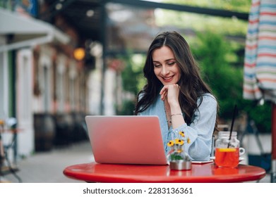 Beautiful woman in casual clothes is using a laptop and smiling while sitting in the cafe outdoors and drinking cold beverage. Chatting online, working outside office. Copy space. - Powered by Shutterstock