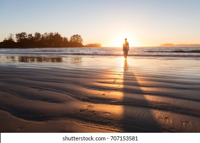 Beautiful woman in a casual dress is walking barefoot on a sandy ocean shore towards the water.  Picture taken on Chesterman Beach, Tofino, Vancouver Island, BC, Canada, during golden summer sunset.