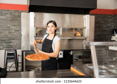 Beautiful Woman Chef Wearing Black Uniform Holding Pizza Peel With Freshly Baked Pepperoni Cheese Pizza In Hand While Placing It On Counter And Looking At Camera In Pizza Shop