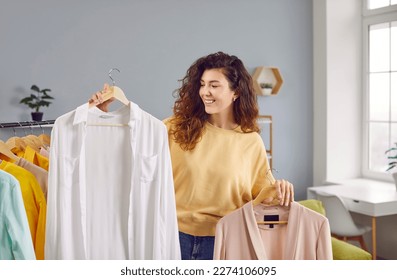 Beautiful woman choosing clothes on rack at home. Attractive smiling young woman holding hangers with shirts, thinking what to wear. Girl trying to choose outfit dressing - Powered by Shutterstock