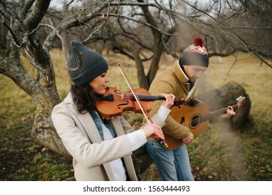 Beautiful Woman In A Coat Plaing Violin With Her Eyes Closed. Next To Her A Warmly Dressed Man Playing Guitar Without Strap, Leaning Forward. Both Wearing Watch Caps. Leafless Trees All Around Them.