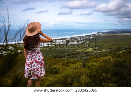 beautiful woman in colorful dress and elegant hat watching panorama of coolum beach and sunshine coast from the top of mount coolum, queensland, australia	