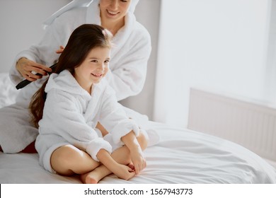 Beautiful Woman Combing Hair To Her Daughter After Shower, Sitting On Bed In Light Bedroom, Wearing Towel And Bathrobe