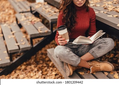 Beautiful woman in cozy outfit reading book and drink coffee sitting on wooden bench in autumn park.