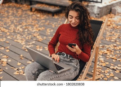 Beautiful woman in cozy outfit works at laptop and using phone while sitting on chair near cafe in autumn park.