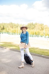 Beautiful Woman Cross Empty Road With Cardboard Poster And Suitcase. Lady In Hat And Denim Outfit Escape From City By Auto Stop To Go Anywhere. Travelling, Freedom, Hitchhiking, Vacation.