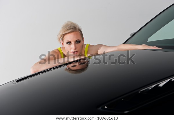 Beautiful woman\
crouching behind her car as she rests her chin while peering over\
the hood with her\
reflection.