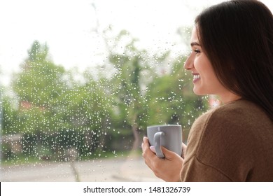 Beautiful Woman With Cup Of Coffee Smiling Near Window Indoors On Rainy Day