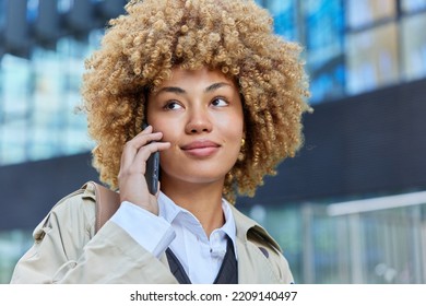 Beautiful woman with curly blonde hair has telephone conversation focused somewhere enjoys pleasant talk dressed in stylish outerwear poses outdoors against blurred background. Technology concept - Powered by Shutterstock