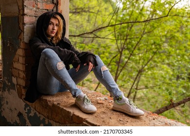 A beautiful woman with curly brown hair in rock Gothic grunge style: torn jeans, black mantle with hood, sitting on the broken window of an abandoned industrial building with brick walls. Depression - Powered by Shutterstock