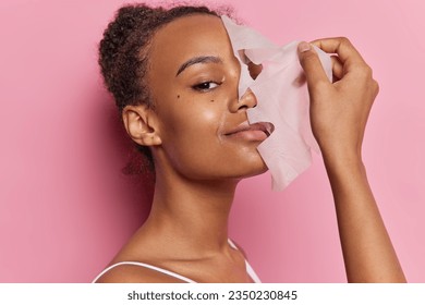 Beautiful woman with curly combed hair puts on facial mask for skin treatment smiles gently wears t shirt isolated over pink wall. Young female model removing from face skincare hydrating sheet mask - Powered by Shutterstock