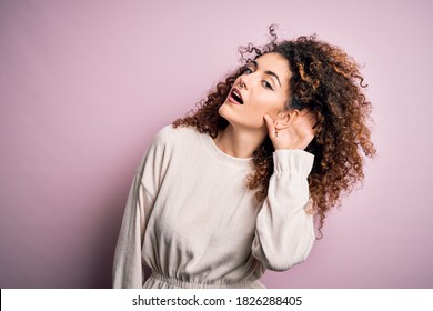 Beautiful Woman With Curly Hair And Piercing Wearing Casual Sweater Over Pink Background Smiling With Hand Over Ear Listening An Hearing To Rumor Or Gossip. Deafness Concept.