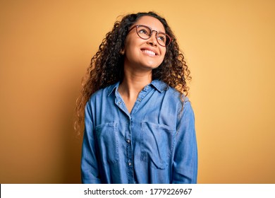 Beautiful woman with curly hair wearing casual denim shirt and glasses over yellow background looking away to side with smile on face, natural expression. Laughing confident.