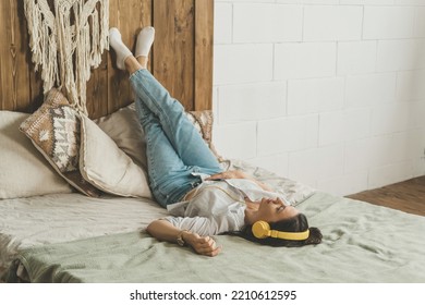 A beautiful woman with dark hair is lying on the bed at home in headphones. A woman rests and relaxes after a working day. The girl got tired and took a break. - Powered by Shutterstock