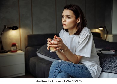 Beautiful woman with dark hair and a white T-shirt sits thoughtfully on the bed and holds a cup of coffee in her hand
