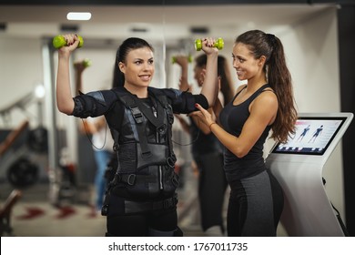 A beautiful woman is doing exercises with female personal trainer in the gym, dressed in a black suit with an EMS electronic simulator to stimulate their muscles.