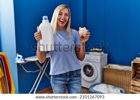 Beautiful woman doing laundry holding detergent bottle and piggy bank smiling and laughing hard out loud because funny crazy joke. 