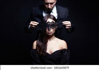 beautiful woman in dress and mask in the hands of man in a suit. Couple over black background. Adorable, elegant girl seducing her handsome boyfriend