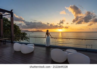 Beautiful woman in dress standing and enjoying the large balcony on sunset with sea view. Back view - Shutterstock ID 2122649228