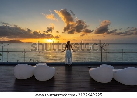 Beautiful woman in dress standing and enjoying the large balcony on sunset with sea view. Back view