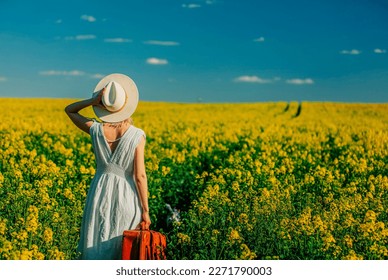 Beautiful woman in dress with suitcase in rapeseed field in spring time 