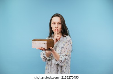 Beautiful woman in a dress and with a wooden box of jewelry in these stands on a blue background, looks at the camera and shows a gesture of silence touching his finger to his lips