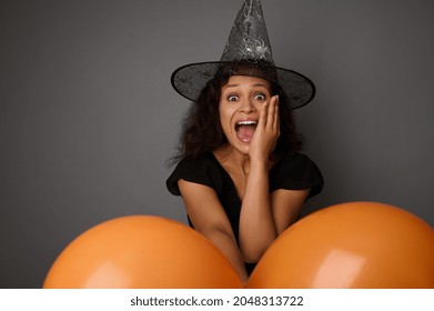 Beautiful woman dressed in black and wizard hat, looks at camera, holds colorful orange air balloons and expresses stupefaction and surprise. Halloween concept on gray background with copy space