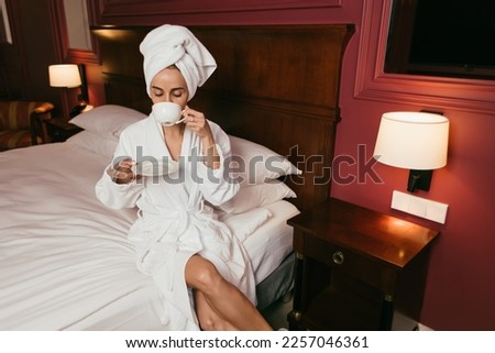 beautiful woman drinking coffee in a white robe and with a white towel on her head, in a hotel room, white bed, white pillows in the bedroom, sunny morning, sun rays