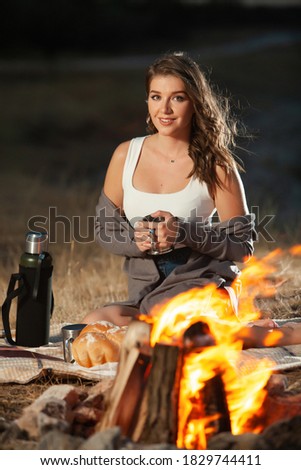 Beautiful woman drinking tea from a thermos by the fire in a romantic nature atmosphere.