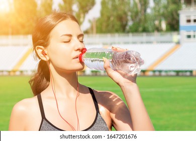 Beautiful Woman Is Drinking Water And Listening To The Music On Headphones At The Stadium. Girl Is Having A Break After Training.