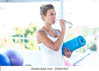 Beautiful Woman Drinking Water While Holding Yoga Mat In Fitness Studio