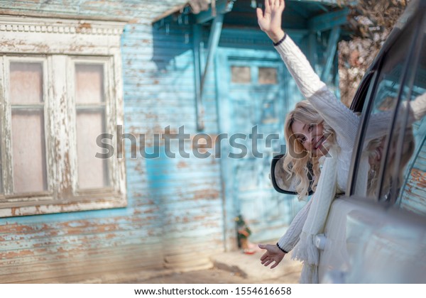 Beautiful\
woman drive the white car and leaning from window of vehicle\
against blue wall of the log village\
house