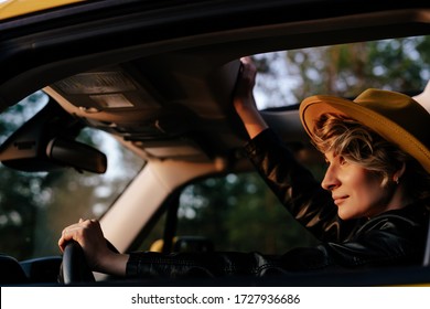 Beautiful Woman Driving Car Side View Portrait. Gorgeous Stylish Blonde Female Sitting At Wheel Watching Sunset Road. Carefree Caucasian Girl Enjoying Comfortable Auto Journey With Open Sunroof