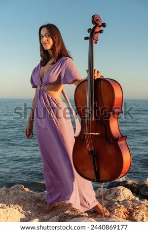 beautiful woman in an elegant purple dress posing on some rocks holding a large musical instrument, bathed in the evening light with the sea in the background