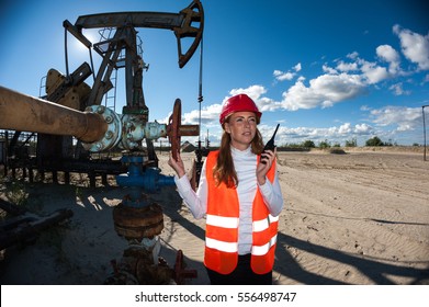 Beautiful woman engineer in the oilfield wearing red helmet and work clothes. Pump jack and wellhead valve background. Oil and gas concept.