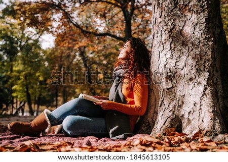 A beautiful woman enjoying the sun in a park in an autumnal day. She is sitting and leaning against a tree while having a book on her legs. Lifestyle autumnal outdoors