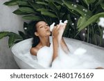 A beautiful woman enjoys a playful bubble bath in a tropical outdoor setting, surrounded by lush greenery. Ideal for concepts of relaxation, luxury, and spa lifestyle in advertisements and promotional