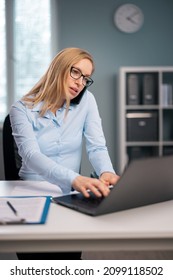 Beautiful Woman In Eyeglasses And Formal Clothes Typing On Modern Laptop And Talking On Mobile. Busy Mature Lady Working At Modern Bright Office.